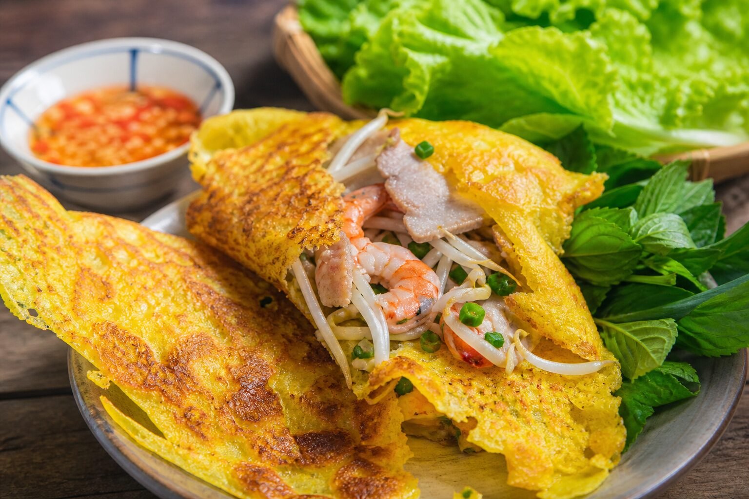 A close-up serving of Vietnamese banh xeo opened to show shrimp, pork, bean sprouts, herbs, and dipping sauce.