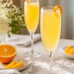 Pale golden French mimosa cocktail in a sparkling flute with an orange twist at a brunch table.