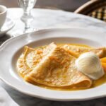 French crepes Suzette folded in glossy orange butter sauce with orange zest and vanilla ice cream.