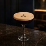 An Espresso Martini with dark coffee cocktail, thick tan crema, and three coffee beans in a coupe glass.