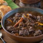 A bowl of glossy Guyanese pepperpot beef stew with bread on an earthy wooden table.