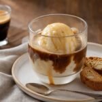 A scoop of vanilla gelato covered with espresso in a glass cup beside crisp almond biscotti.