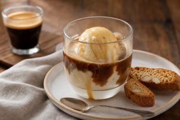 A scoop of vanilla gelato covered with espresso in a glass cup beside crisp almond biscotti.