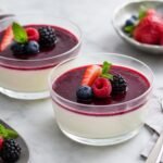 Silky Italian panna cotta topped with berry sauce and fresh berries on a pale dessert plate.