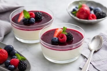 Silky Italian panna cotta topped with berry sauce and fresh berries on a pale dessert plate.