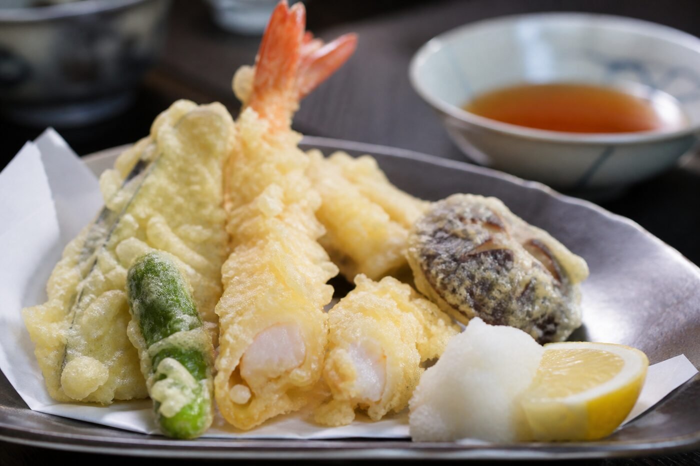 Close-up of airy crisp tempura shrimp and vegetables with dipping sauce and grated daikon.