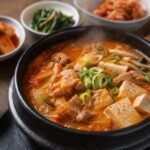 Steaming Korean kimchi jjigae with kimchi, tofu, pork, scallions, and mushrooms in a black stone bowl.