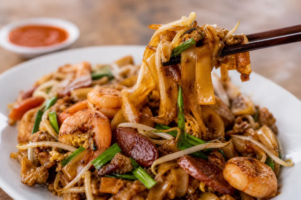 A close-up serving of char kway teow with flat rice noodles, shrimp, Chinese sausage, egg, bean sprouts, and scallions.