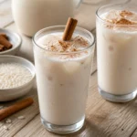 Two glasses of Mexican horchata over ice with cinnamon, rice, and a pitcher in the background.