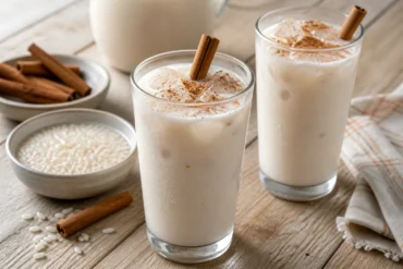 Two glasses of Mexican horchata over ice with cinnamon, rice, and a pitcher in the background.