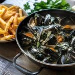 Belgian moules-frites with steamed mussels in creamy white wine broth beside crisp golden fries and parsley.