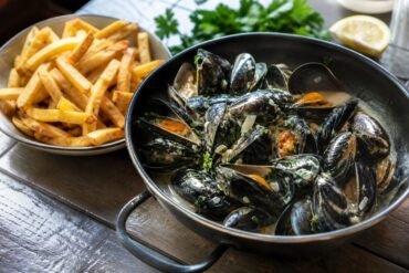 Belgian moules-frites with steamed mussels in creamy white wine broth beside crisp golden fries and parsley.