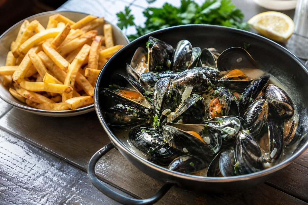 Belgian moules-frites with steamed mussels in creamy white wine broth beside crisp golden fries and parsley.