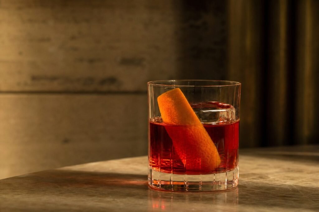 A ruby-red Negroni cocktail with clear ice and a wide orange peel in a rocks glass.
