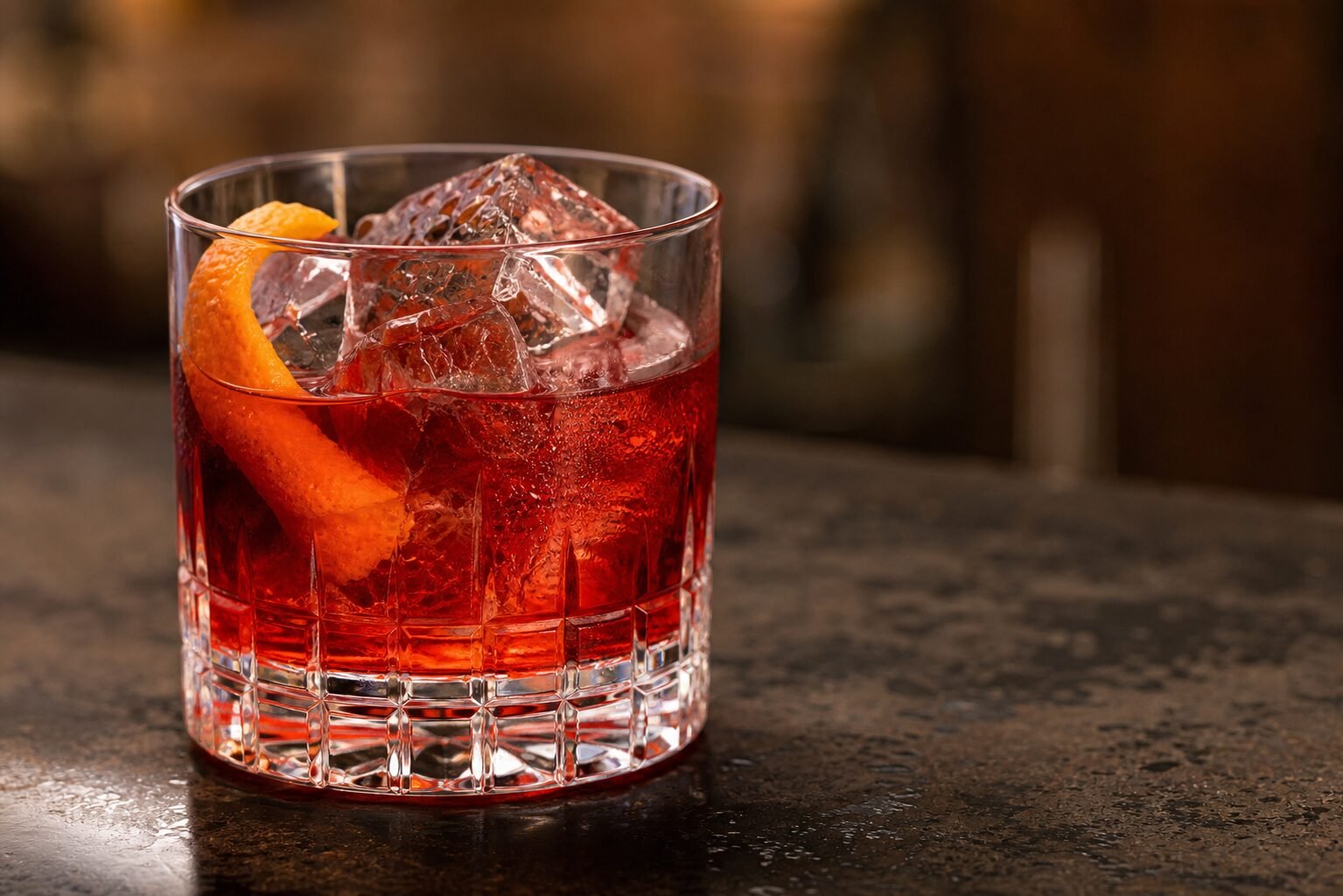 Close view of a chilled Negroni with red cocktail, clear ice, condensation, and orange peel.