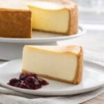 A tall slice of classic New York cheesecake with creamy filling, graham cracker crust, and berry compote on the side.