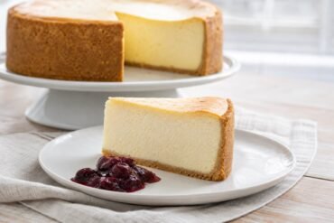 A tall slice of classic New York cheesecake with creamy filling, graham cracker crust, and berry compote on the side.