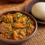 A bowl of Nigerian egusi soup with beef, leafy greens, red palm oil, and pounded yam.