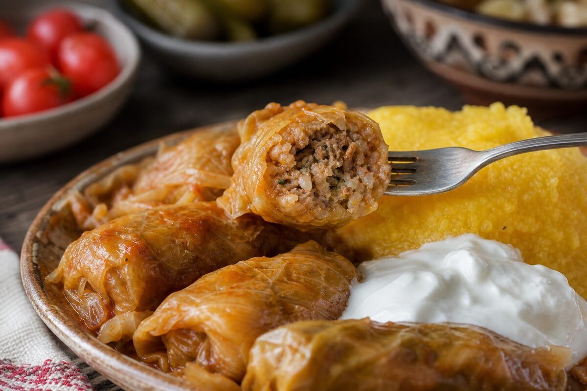 Close serving of Romanian sarmale showing a tender cabbage roll filled with pork and rice beside sour cream and polenta.