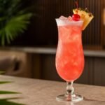 A rosy Singapore Sling cocktail with ice, light foam, pineapple, and a cherry.