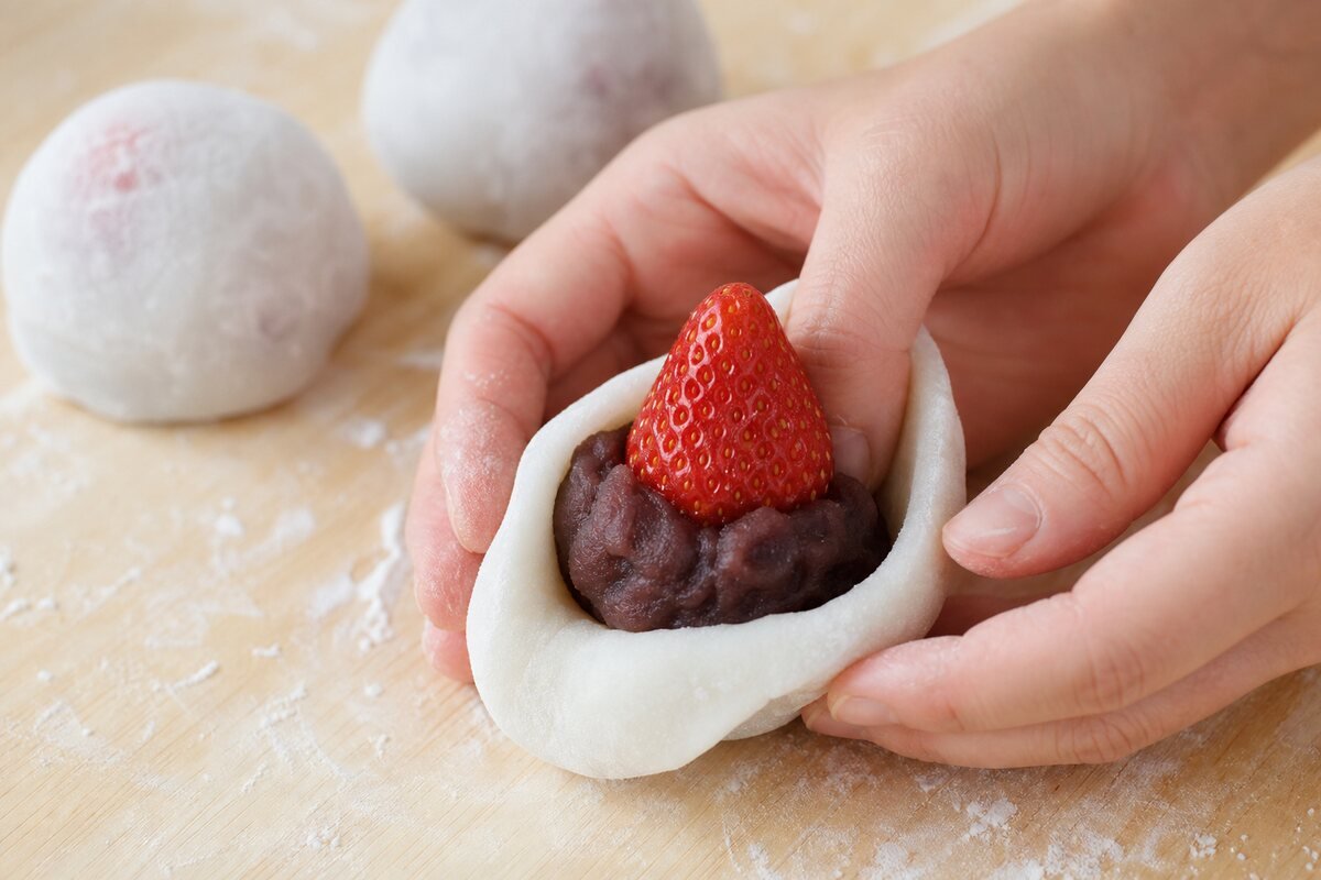 Hands wrap soft mochi dough around a strawberry covered with sweet red bean paste.