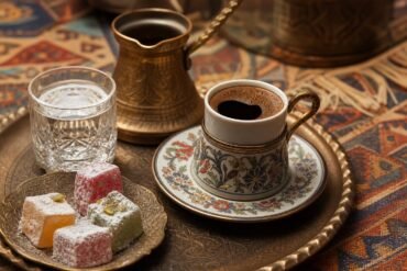 Turkish coffee in a traditional cup with foam, Turkish delight, a brass coffee pot, and water.