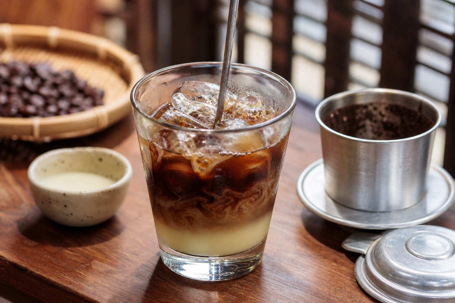 Vietnamese iced coffee stirred over ice with sweetened condensed milk and a metal phin filter nearby.