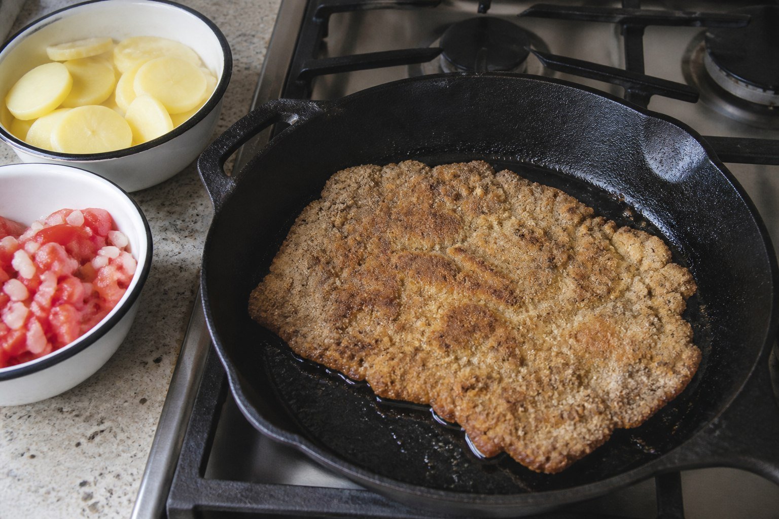 A thin breaded silpancho beef cutlet frying beside sliced potatoes and tomato salad.