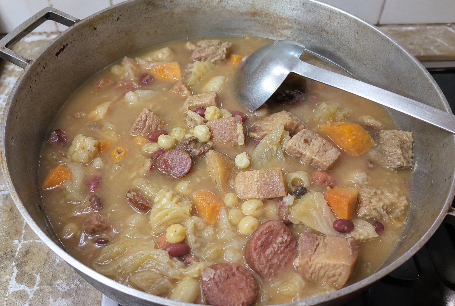 Cachupa simmering in a seasoned metal pot with pork, sausage, beans, corn, and vegetables.