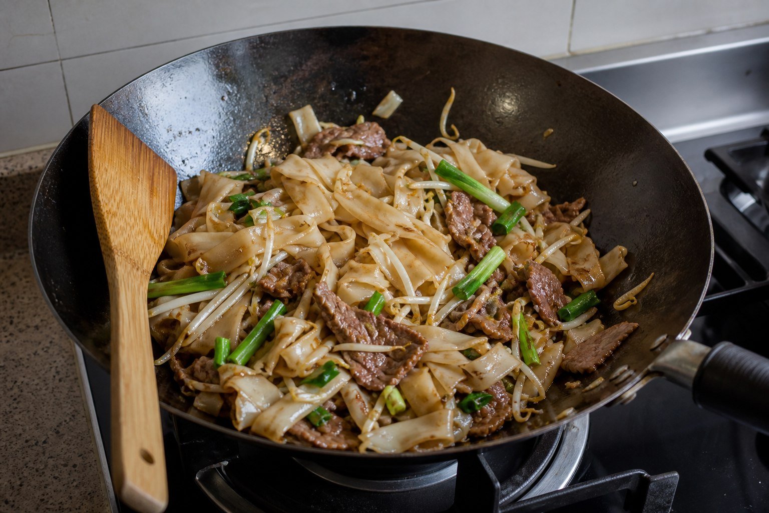 Broad rice noodles, beef, sprouts, and scallions being stir-fried in a carbon-steel wok.