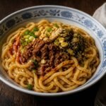 A bowl of Sichuan dan dan noodles topped with pork, greens, peanuts, and chili oil.