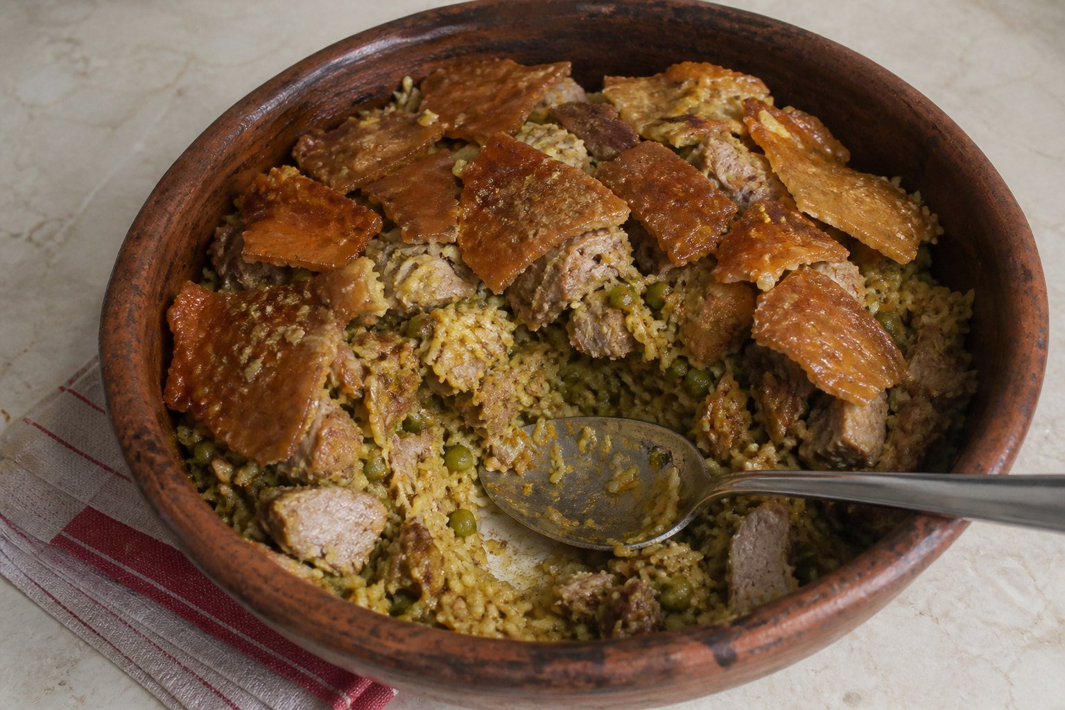 Rustic baking dish of Colombian lechona with rice, peas, pork, crisp skin, and serving spoon.