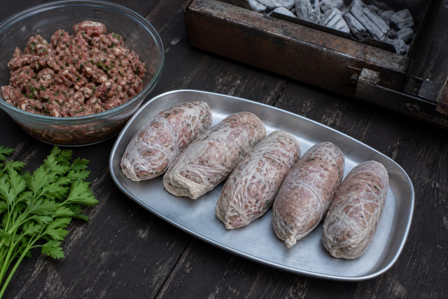 Raw sheftalia wrapped in caul fat beside seasoned minced meat and a charcoal grill.