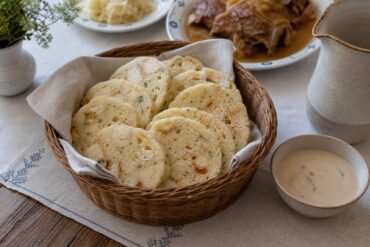 Sliced Czech bread dumplings, houskové knedlíky, stacked in a basket beside creamy sauce.