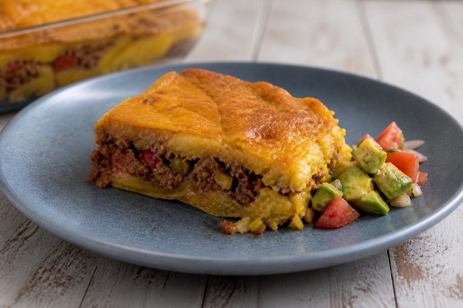 A slice of Dominican pastelón served with avocado and tomato salad.