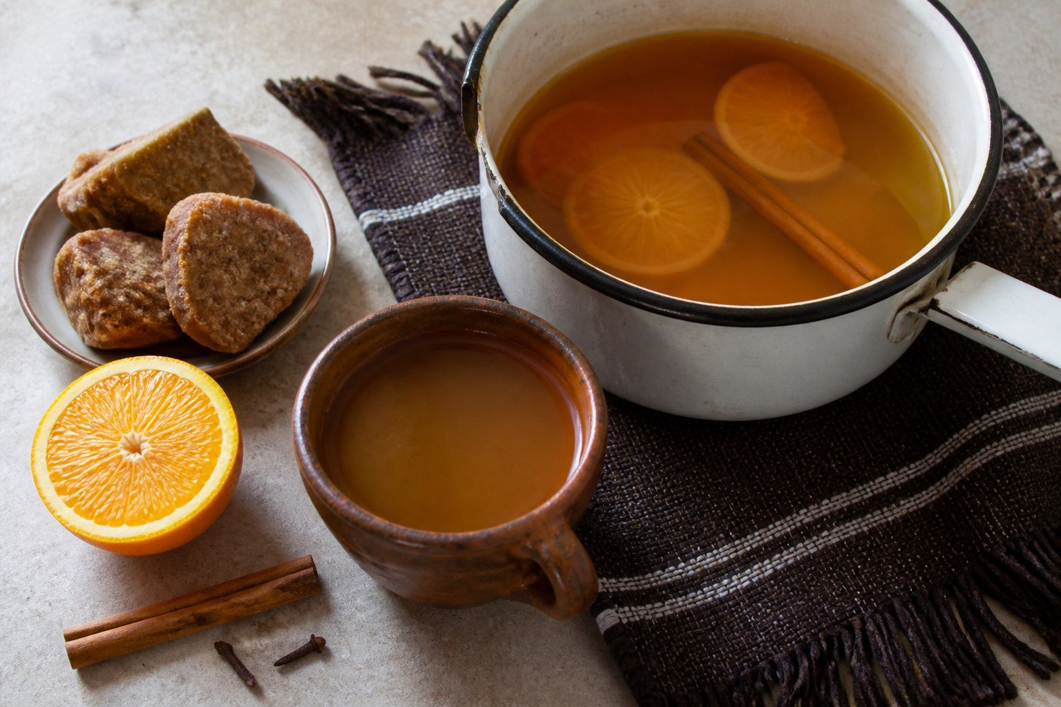 Ecuadorian canelazo in a clay cup beside an enamel pot, panela, orange, cinnamon, and cloves.