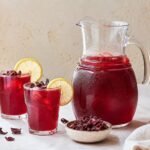 Ruby-red Egyptian hibiscus tea served in glasses beside a pitcher and dried petals.