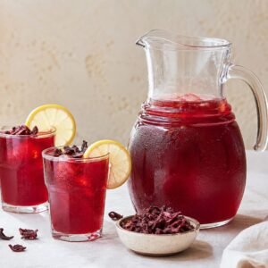 Ruby-red Egyptian hibiscus tea served in glasses beside a pitcher and dried petals.