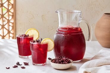 Ruby-red Egyptian hibiscus tea served in glasses beside a pitcher and dried petals.