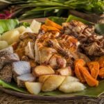 Fijian lovo with roasted chicken, pork, cassava, taro, and sweet potato arranged on banana leaves.