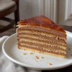 Slice of Hungarian Dobos torte with golden sponge, chocolate buttercream, and caramel top.