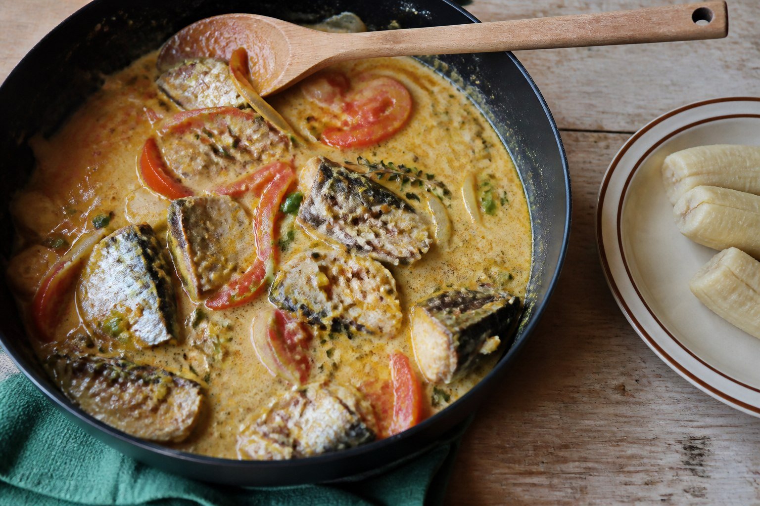 Mackerel rundown simmering in a skillet beside boiled green banana.