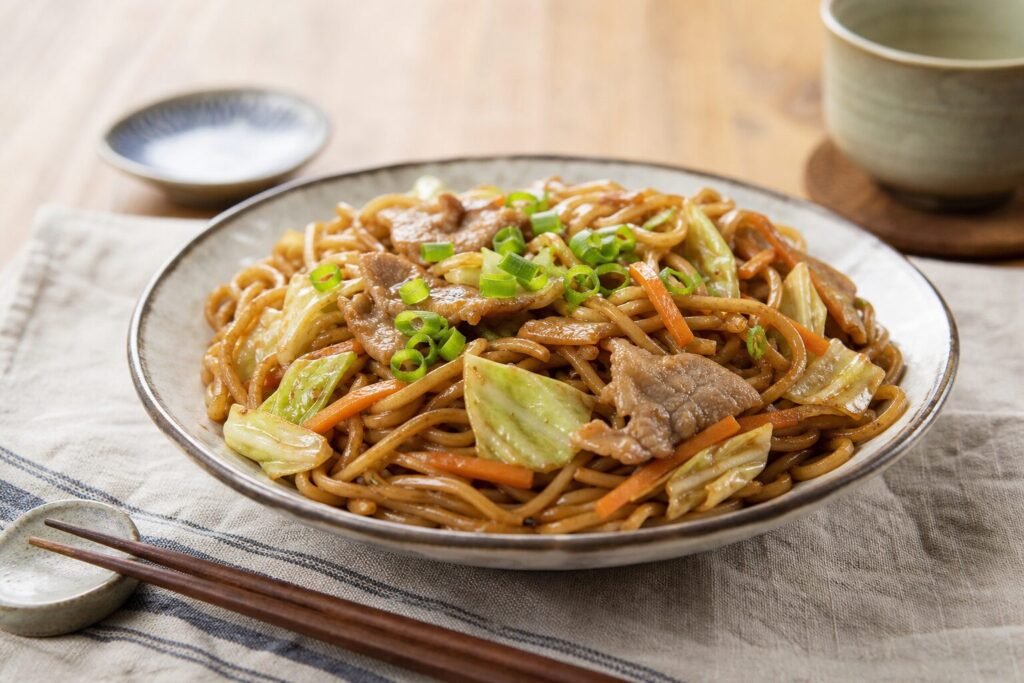 Glossy Japanese yakisoba noodles with pork, cabbage, carrots, and green onions in a ceramic bowl.