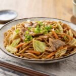 Glossy Japanese yakisoba noodles with pork, cabbage, carrots, and green onions in a ceramic bowl.