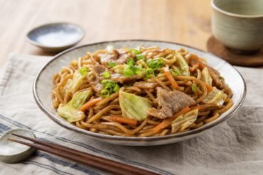 Glossy Japanese yakisoba noodles with pork, cabbage, carrots, and green onions in a ceramic bowl.