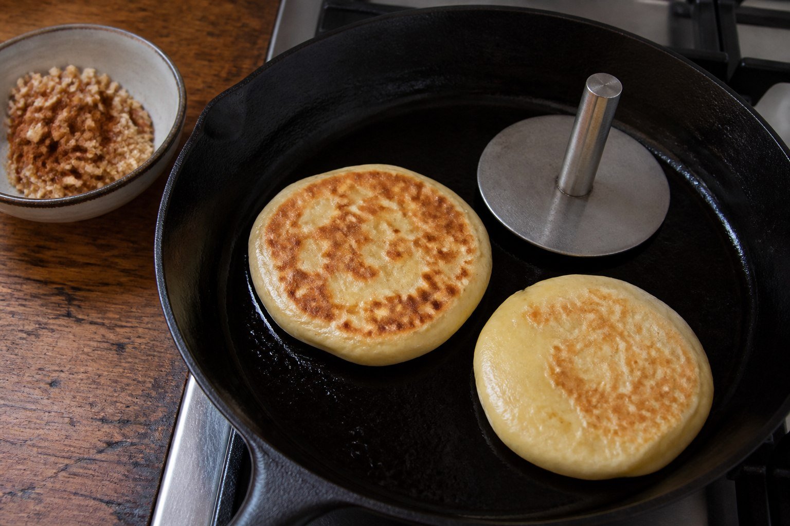 Korean hotteok pancakes cooking in a cast-iron skillet beside a metal press and filling.
