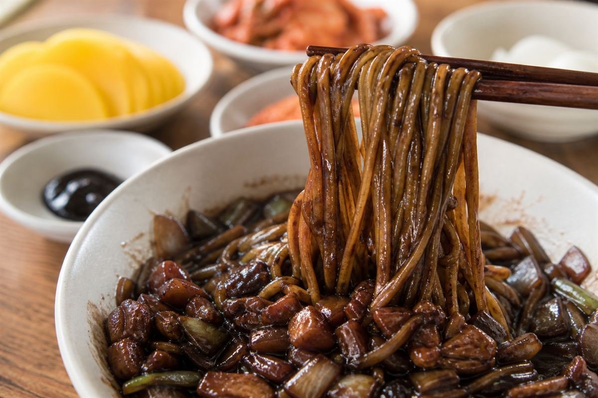 Springy Korean jajangmyeon noodles lifted with chopsticks beside pickled radish.