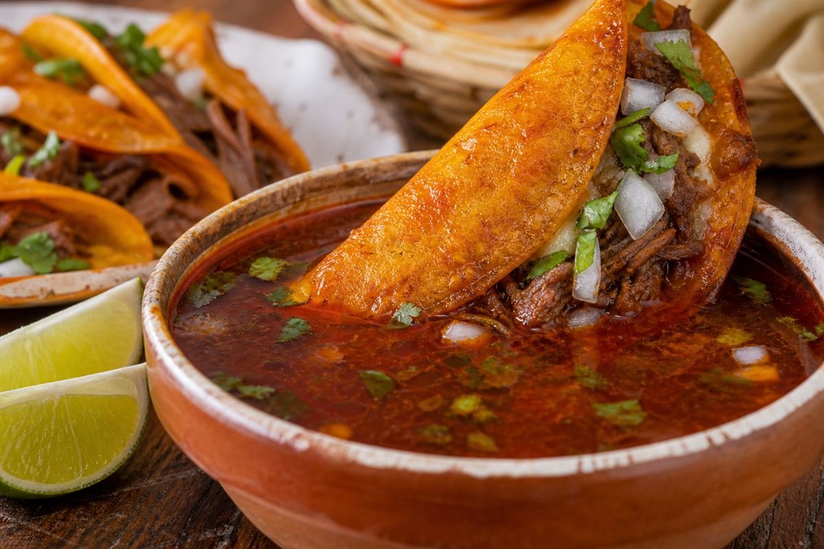 A birria taco being dipped into rich red consommé with cilantro and lime.
