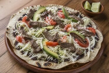 An open Oaxacan tlayuda with black beans, quesillo, cabbage, avocado, and grilled beef.