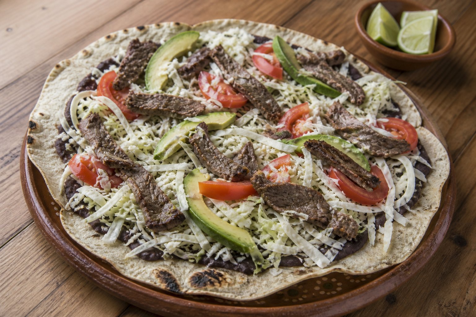 An open Oaxacan tlayuda with black beans, quesillo, cabbage, avocado, and grilled beef.